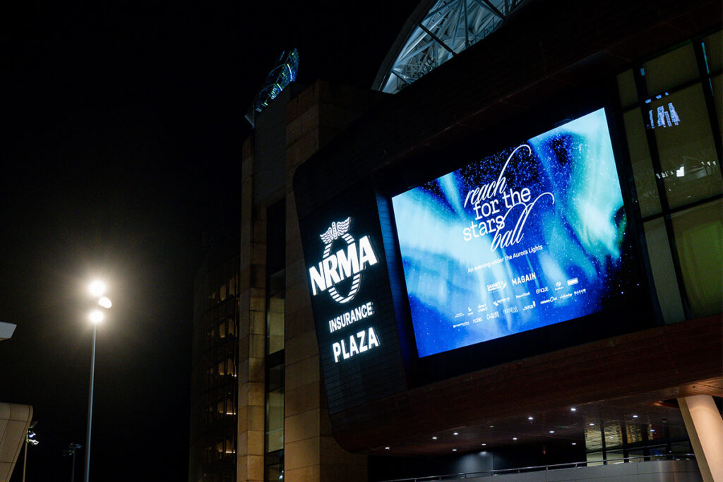 Large digital screen outside NRMA Insurance Plaza at night promoting the Reach for the Stars Ball, An evening under the Aurora Lights.