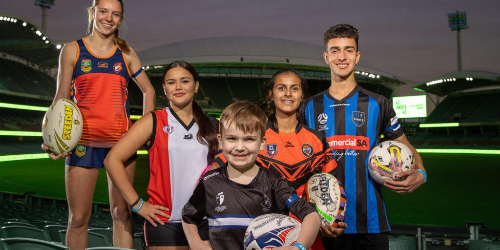 Five young athletes in rugby league, soccer and football club jerseys smiling with their balls inside a stadium at dusk.